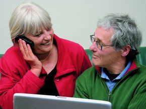 Cuplu în vârstă, în fața laptop-ului, uitându-se unul la celălalt, în timp ce ea vorbește la telefon
